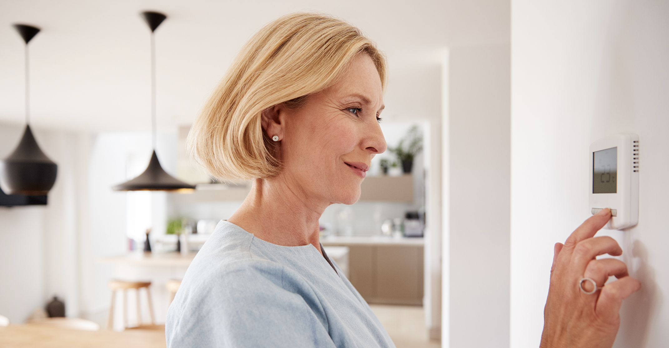 A medium-long, horizontal photograph captures a middle-aged woman with a short blonde bob smiling as she looks at a white digital thermostat on a wall. She is on the right side of the frame, from her right profile, dressed in a light-blue linen top. Her right index finger is pressing a button on the bottom right of the thermostat, which is mounted on a white wall. The thermostat has a grey display screen with small numbers and text. Her left hand, wearing a large, geometric silver ring on her middle finger, rests near her. The background on the left features a modern, open-plan kitchen and dining area, including blurred black pendant lights, a light wood counter with stools, beige cabinets, and plants on a shelf. The room is well-lit with natural light. The focus is on the woman and the thermostat.