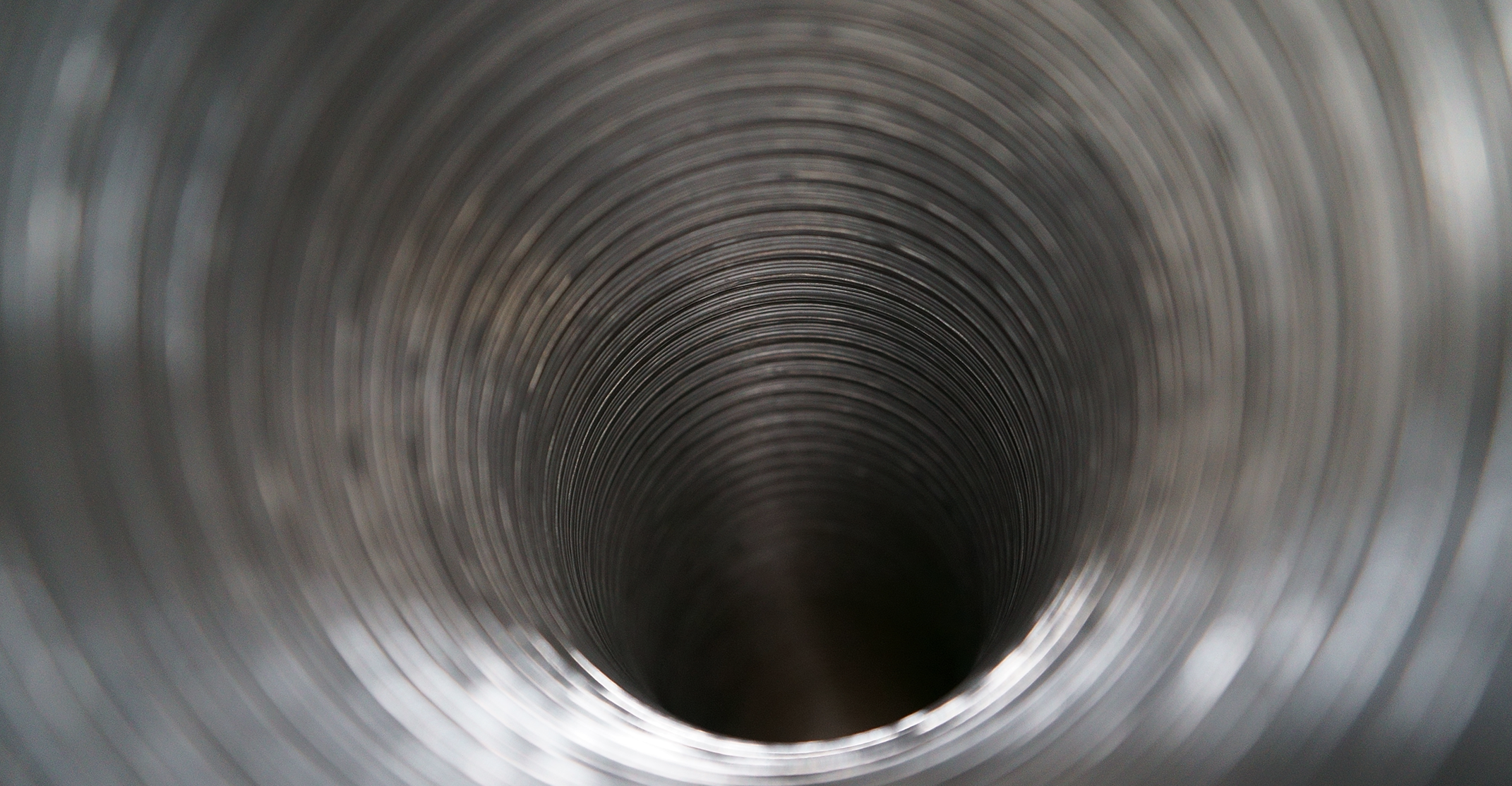 An abstract, close-up photograph looking directly through the center of a shiny, flexible metallic duct or pipe. The interior is characterized by a series of repeating, concentric silver rings that create a deep tunnel effect, leading to a dark, shadowy center. The metal has a brushed or polished finish that reflects soft light, creating bright highlights along the ridges of the coils. The perspective creates a sense of depth and symmetry, with the outer rings appearing larger and slightly out of focus, while the inner rings draw the eye toward the darkness at the end of the tube.