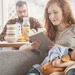 Woman with long red hair sitting on a sofa reading a book, wearing a light-colored long-sleeve top, while a young child sleeps beside her with their head resting on a cushion and holding a soft toy. The child is dressed in a blue shirt and is partially covered by a textured blanket. In the background, a man wearing glasses and a checkered shirt sits at a dining table with breakfast items, including a teapot, cups, a glass bottle of juice, and a bowl, with flowers placed on the table. Natural light enters through windows, illuminating the living and dining area.