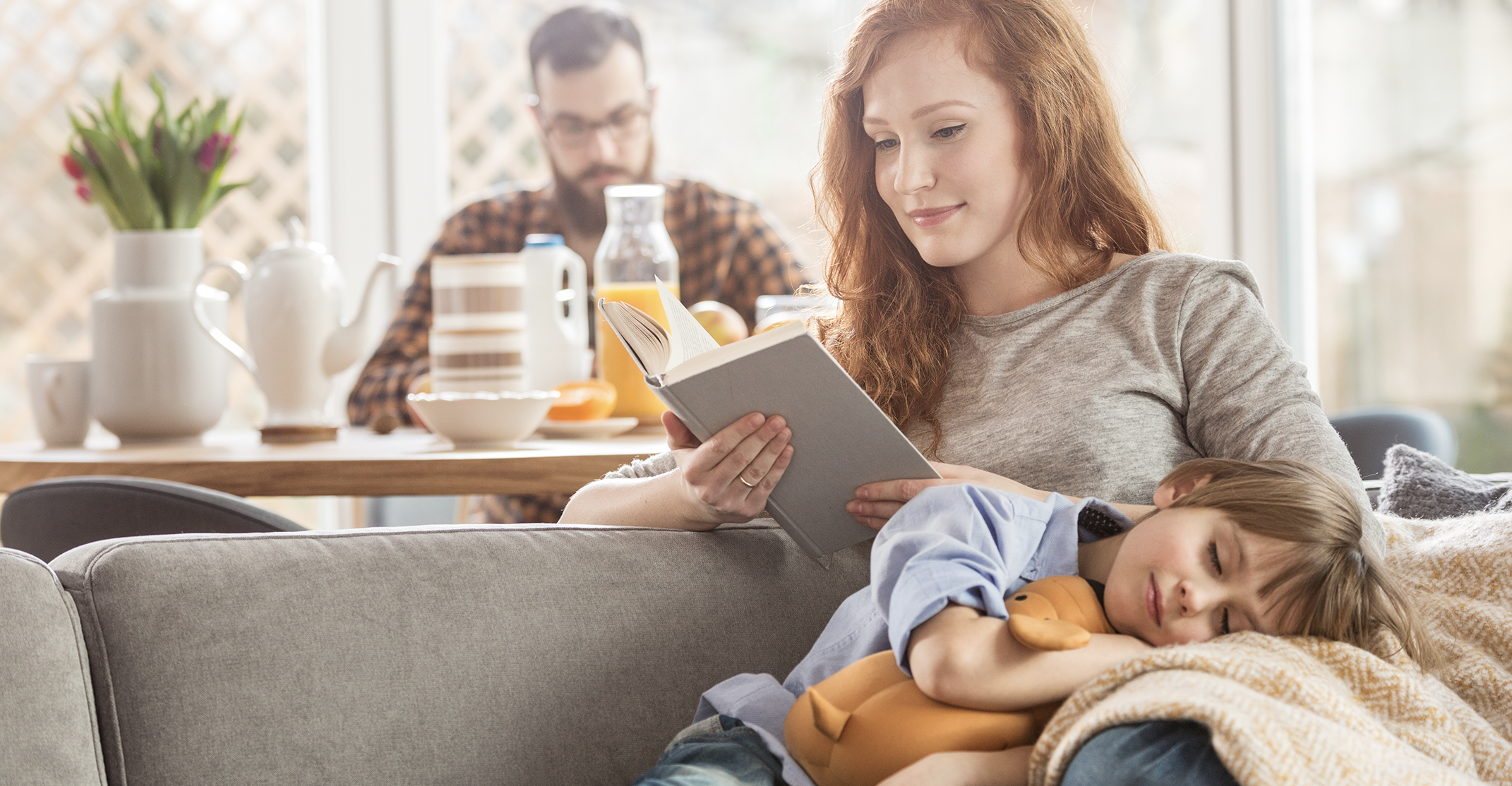 Woman with long red hair sitting on a sofa reading a book, wearing a light-colored long-sleeve top, while a young child sleeps beside her with their head resting on a cushion and holding a soft toy. The child is dressed in a blue shirt and is partially covered by a textured blanket. In the background, a man wearing glasses and a checkered shirt sits at a dining table with breakfast items, including a teapot, cups, a glass bottle of juice, and a bowl, with flowers placed on the table. Natural light enters through windows, illuminating the living and dining area.