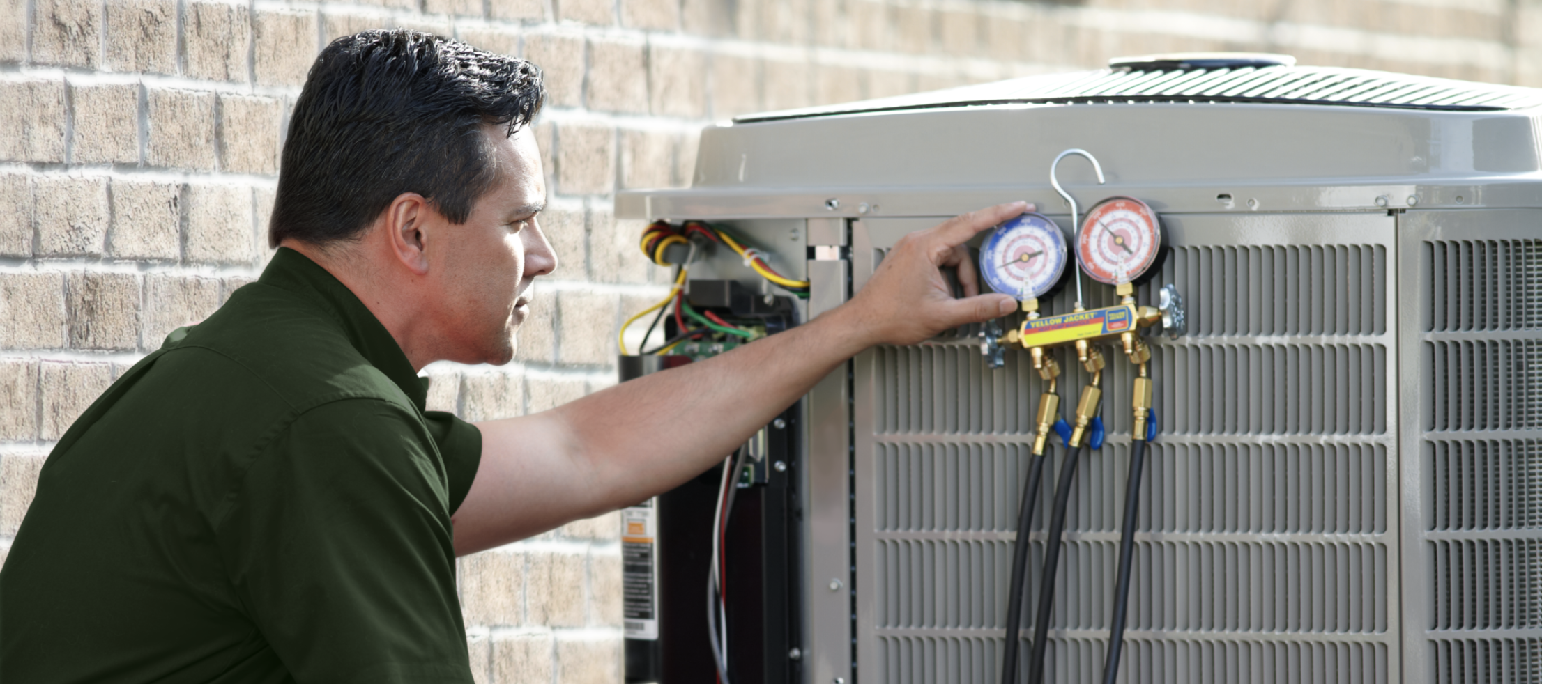 A medium-long, horizontal photograph captures a male HVAC technician inspecting an outdoor air conditioning condenser unit against a light-colored brick wall. The technician, seen from the waist up in a side profile on the right, wears a dark olive-green button-down shirt. He is looking intently at a yellow manifold gauge set hooked onto the unit's metal grille. His right index finger points to the blue low-pressure gauge on the left. Several black hoses extend from the bottom of the gauge set. The scene is illuminated by soft, natural sunlight from the upper right, and the focus is sharp on the technician's focused expression and the diagnostic tools.