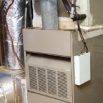 A medium-long, horizontal photograph of a beige gas furnace with exposed metal ductwork is shown against a grey tiled and white concrete block wall. The furnace unit is on the right, and the ductwork is on the left. The front of the furnace is beige, with horizontal vents and a central slot. It has a vertical grey exhaust pipe and various wires and pipes running to it. Below the main vents, the lower panel of the furnace is beige and has diagonal vents. The ductwork on the left is mostly silver and foil-covered, and is connected to the furnace with multiple angles and elbows. A large vertical section of the silver ductwork is in the center-left. The wall behind the furnace is beige at the top and light grey below. The background includes a wooden post and part of a grey concrete block wall. On the floor to the left, a small green electronic board is visible on a yellow board. The overall image is slightly messy and industrial.