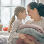 A tender and heartwarming moment is shared between a mother and her young daughter in a softly lit, cozy indoor setting. Both are dressed in warm, knitted sweaters, suggesting a cool or chilly environment. The mother gently holds a stack of folded blankets in soft pastel tones—pink and gray—cradled in her arms, while the little girl leans in close, smiling brightly. Their faces are nearly touching as they gaze into each other’s eyes with affection and joy, creating a strong sense of emotional connection. The child’s hair is neatly tied back, and her expression reflects happiness and comfort. In the background, a large window allows natural light to fill the room, and a basket filled with colorful yarn or fabric sits nearby, adding to the warm, homely atmosphere.