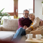 An elderly couple sits close together on a light-colored sofa in a cozy living room. The man, wearing glasses, a burgundy sweater, and blue pants, has his arm wrapped around the woman beside him. The woman, with short gray hair, wears a beige cardigan over a yellow top and leans her head gently against his shoulder while smiling. Their hands rest together on their laps. Behind them, large windows let in natural light, and several green houseplants are placed around the room. Soft decorative lights are visible on a shelf in the background. In the foreground, a small table holds two white cups, slightly out of focus. The space appears warm and neatly arranged with neutral tones and simple furnishings.