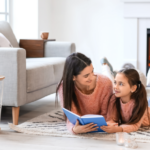 A horizontal, medium-long shot of a young woman and a young girl lying on their stomachs on a thick, grey, woven rug in a brightly lit living room. Both are wearing matching fuzzy, coral-pink sweaters. The woman, on the left with long dark hair, holds an open blue book and smiles down at it. The young girl, on the right, looks up at the woman with a slight smile. In the background to the right, a white modern fireplace contains a glowing orange electric fire. To the left, a grey armchair, a small wooden side table with logs underneath, and a window with natural light are visible. Several small lit candles in glass holders are scattered on the floor around them, adding a warm glow to the cozy scene. The focus is sharp on the two individuals in the foreground.
