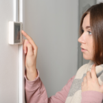 Gemini said A medium close-up, horizontal photograph shows a young woman with shoulder-length brown hair, viewed from her left side. She is wearing a pink sweatshirt and is wrapped in a grey and cream plaid blanket, which she holds tightly around her. She is looking towards the left, at a small, white digital thermostat mounted on the wall. Her left index finger is pressed against the screen of the thermostat, and her expression is one of slight concern. Her hand is placed near her chest, beneath the blanket. The wall is light grey, and a white corner trim is visible. The background is slightly out of focus.