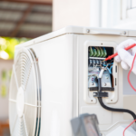 A close-up view of an outdoor air conditioning unit being serviced, with a technician’s gloved hand using a red multimeter probe to test electrical connections inside an open access panel. The unit’s internal wiring, terminals, and components are visible, including neatly arranged colored wires and connection points. The technician appears to be checking voltage or continuity, indicating maintenance or troubleshooting work. The AC unit is mounted outside near a building wall, with part of a fan casing visible on the left. The background is softly blurred, suggesting an outdoor residential setting. The scene highlights professional HVAC inspection and repair in progress.