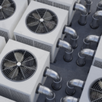 Multiple outdoor HVAC condenser units with square metal housings and large circular fan grilles on top, each unit featuring vented side panels and connected by exposed metallic pipes and curved ductwork that run between the units, with all equipment installed on a flat surface and arranged closely together.