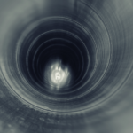 Close-up view inside a cylindrical metal pipe showing smooth interior walls with circular ridges leading toward a darker center. Light reflects along the inner surface, creating a gradient from bright edges to a shadowed opening at the far end, emphasizing the depth and hollow structure of the pipe.