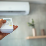 A close-up view of a hand holding a white remote control pointed toward a wall-mounted air conditioner in the background. The remote is in focus, showing several buttons and a small display screen, while the air conditioner unit appears blurred on a light-colored wall. The hand is positioned in the lower left of the image, with fingers wrapped around the remote and the thumb near the buttons. In the background, a small wooden shelf with a potted plant is visible but out of focus, adding depth to the scene. The overall setting appears to be a clean, modern indoor space with soft lighting and neutral tones.