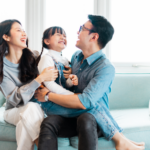 A candid, medium shot of a Japanese family of three – a mother, father, and young daughter – laughing happily together on a light-blue sofa. The mother is on the left, looking to the side with her mouth open in a wide smile, wearing a grey sweater and white pants. The daughter is in the center, sitting in her father’s lap, laughing hard with her eyes closed, wearing denim overalls and a white shirt with pigtails. The father is on the right, looking down at his daughter with a wide smile, wearing a blue button-down shirt and black pants with glasses, holding his daughter tightly around her waist. The background is a bright room with light spilling in from two large windows with sheer curtains. The photograph is well-lit, showing the textures of the family’s clothing and the soft light from the windows. The focus is on the family, with the background slightly blurred.