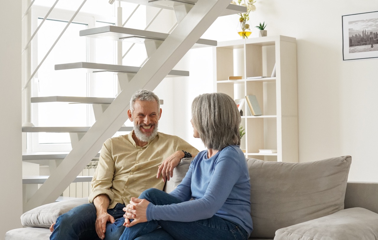 Middle-aged couple sitting on a light-colored sofa in a modern living room, facing each other and smiling while talking. The man, wearing a beige shirt and jeans, leans back with one arm resting on the sofa, while the woman in a blue sweater and jeans sits turned toward him with her hands together. Behind them is a white open staircase with floating steps and a shelving unit holding books and small decor items. Natural light fills the space through large windows, creating a bright, clean interior setting.