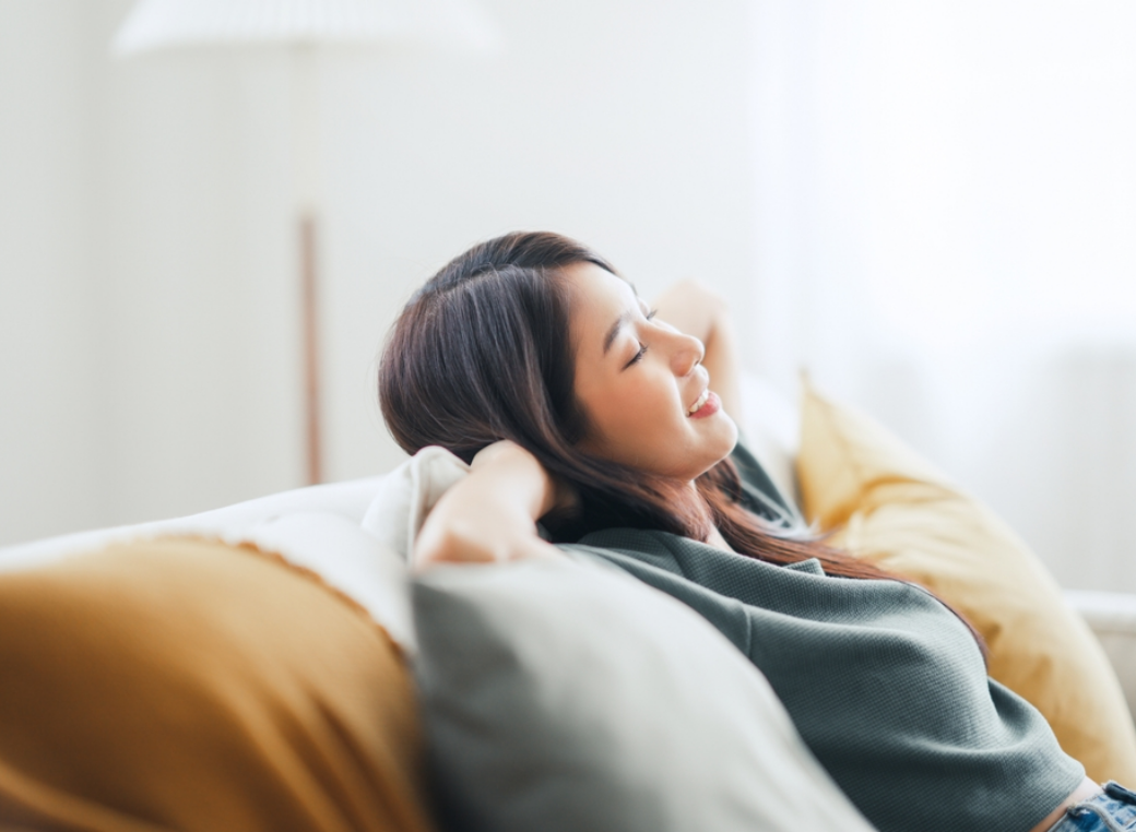 Gemini said A medium-close, right profile photograph captures a young woman with dark brown hair relaxing with her eyes closed on a grey sofa, leaning back against multiple large, plush throw pillows. Her hands are interlaced and positioned behind her head, providing a restful support as she leans against a light-grey, patterned pillow and another in a light-yellow fabric. She wears a grey long-sleeved shirt with a ribbed texture and is in a comfortable, relaxed state, with a small smile on her face. Natural light streams in from a window in the background, creating a bright and airy ambiance. The background is softly out of focus, featuring a blurred standing lamp with a white lampshade and a light-grey wall. The focus is on the woman, with soft light from the window creating a gentle gradient of light on her face and hair.