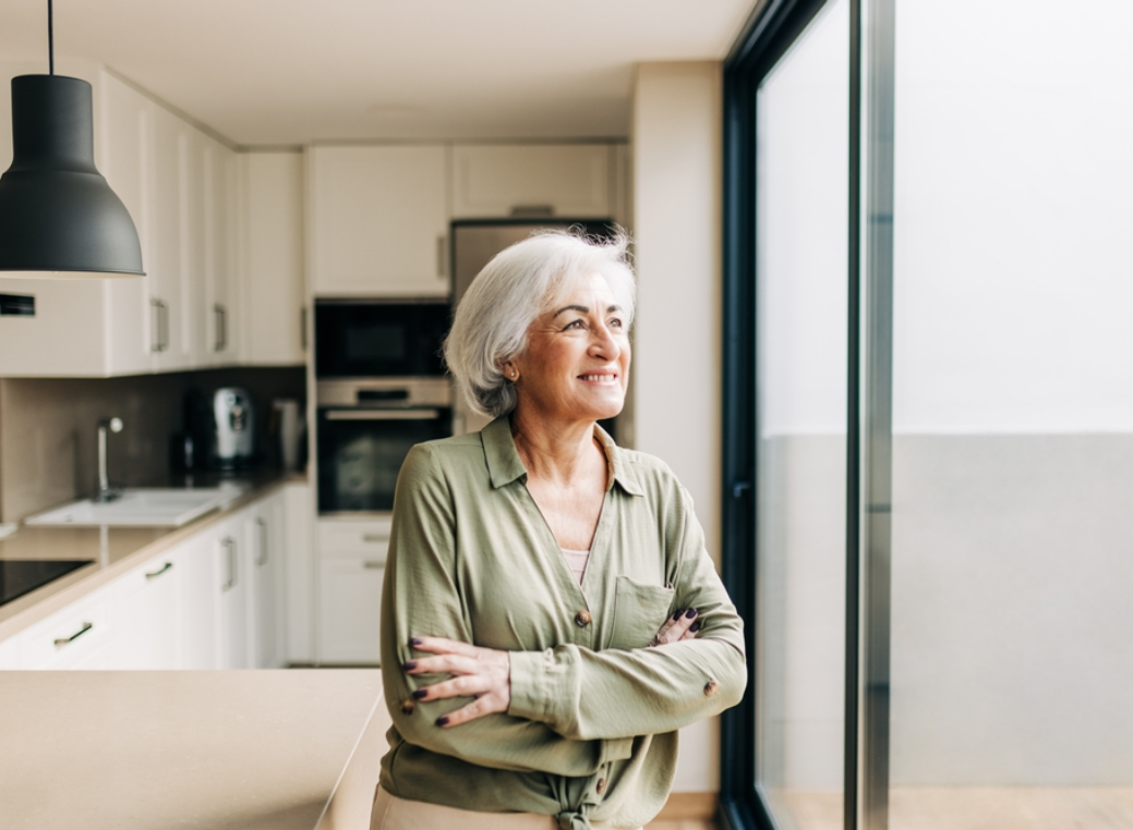An older woman with short gray hair stands in a modern, well-lit kitchen, smiling gently as she looks out through a large glass door or window. She wears a light green button-up blouse with the sleeves rolled down and has her arms comfortably crossed, conveying a relaxed and content posture. The kitchen features clean, minimalist design elements, including white cabinetry, built-in appliances such as an oven and microwave, and a light-colored countertop. Natural light pours in from the glass door beside her, brightening the space and creating a calm, airy atmosphere. The overall scene suggests a peaceful moment of reflection and comfort in a contemporary home setting.