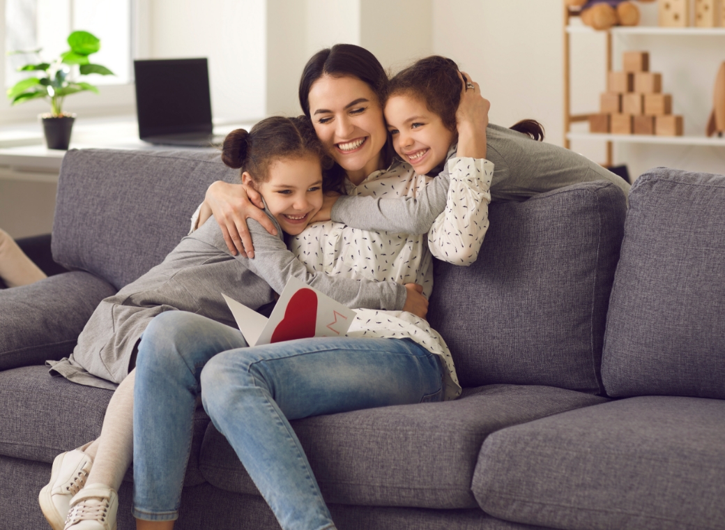 Woman sitting on a gray sofa hugging two young girls who are leaning into her on both sides, all smiling and appearing happy. The girls are wearing casual clothing and have their hair tied back, while the woman wears a light patterned top and jeans. One of the children holds a card with a red heart shape partially visible. The background shows a bright living room with a window, a laptop on a table, and shelves with decorative items.