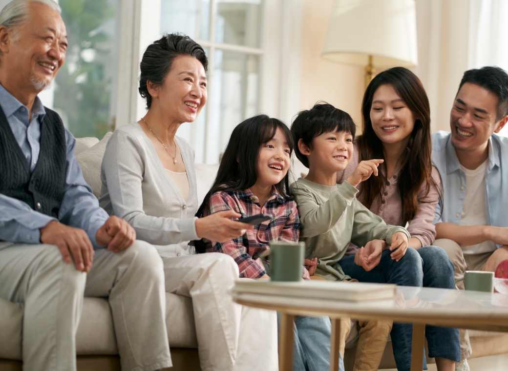 A wide, horizontal photograph shows a multi-generational East Asian family of six sitting together on a grey sofa, laughing and looking toward something off-camera to the right. The family includes a grandfather in a navy vest and light blue shirt, a grandmother in a grey cardigan and pearl necklace holding a black remote, two young children—a girl in a plaid shirt and a boy in an olive shirt—and a younger couple in casual blue and beige clothing. The young boy is pointing excitedly toward the right. In the foreground, a wooden coffee table holds a green mug and a stack of books. The scene is set in a bright, modern living room with soft natural light coming from a window in the background, creating a warm and happy family atmosphere.