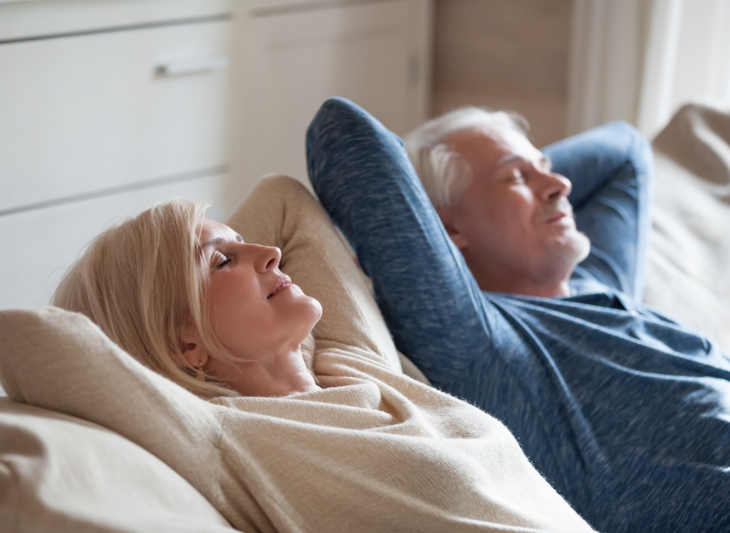 An older couple relaxes comfortably on a sofa in a bright, peaceful living space, both leaning back with their hands resting behind their heads and eyes gently closed. The woman, wearing a soft beige sweater, and the man, dressed in a blue long-sleeve shirt, appear calm and content, enjoying a quiet moment of rest together. Natural light filters into the room, highlighting their relaxed expressions and creating a warm, serene atmosphere that suggests comfort, ease, and a stress-free home environment.