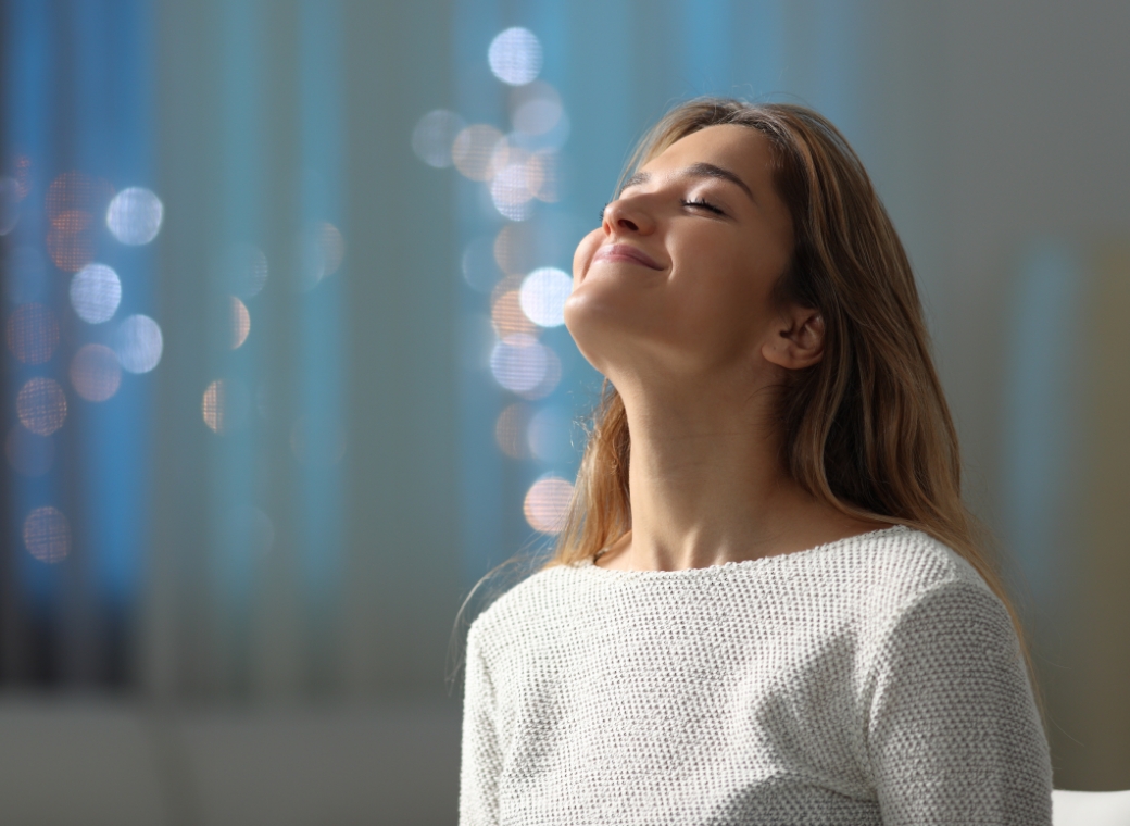Close-up of a woman with long light brown hair wearing a light knit sweater, standing indoors with her eyes closed and head slightly tilted upward. She has a relaxed expression with a gentle smile, as if enjoying fresh air or a comfortable indoor environment. The background is softly blurred with cool-toned lighting and circular bokeh highlights.