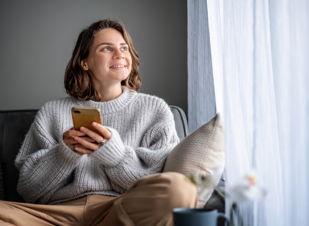 A young woman sits comfortably on a sofa near a window, holding a smartphone in both hands while gazing upward with a soft, content smile. She wears a cozy gray knit sweater and relaxed-fit pants, suggesting a casual, at-home setting. Natural light filters through sheer curtains beside her, illuminating her face and creating a calm, reflective mood. A cushion rests behind her for support, and a cup and small plant are visible nearby, adding to the warm, peaceful atmosphere of a quiet moment spent indoors.