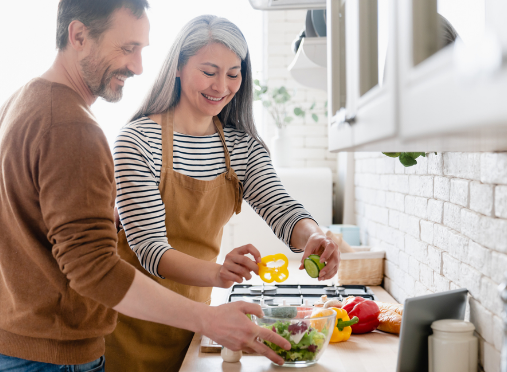 Man and woman standing together in a bright kitchen preparing food, both smiling as they add sliced vegetables to a glass bowl on the countertop. The woman, wearing a striped long-sleeve shirt and a brown apron, holds slices of yellow bell pepper and cucumber over the bowl, while the man in a brown sweater stands beside her assisting. Fresh ingredients including red bell peppers, carrots, and greens are spread across the counter. The background features white cabinets, a tiled backsplash, and natural light illuminating the space.