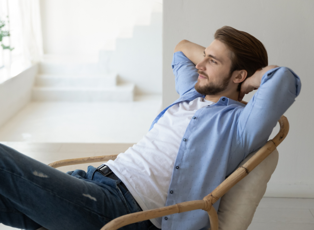 A wide, horizontal shot captures a relaxed young man with a beard and brown hair reclining in a bamboo lounge chair. He is shown from a side profile, leaning back with both hands behind his head and a contented smile as he looks toward the right. He wears a light-blue button-down shirt over a white t-shirt, paired with distressed blue jeans and a black belt. The setting is a bright, minimalist interior with white walls and a staircase in the softly blurred background, suggesting a peaceful home environment. Natural light from a nearby window illuminates the scene, creating a clean and airy atmosphere.