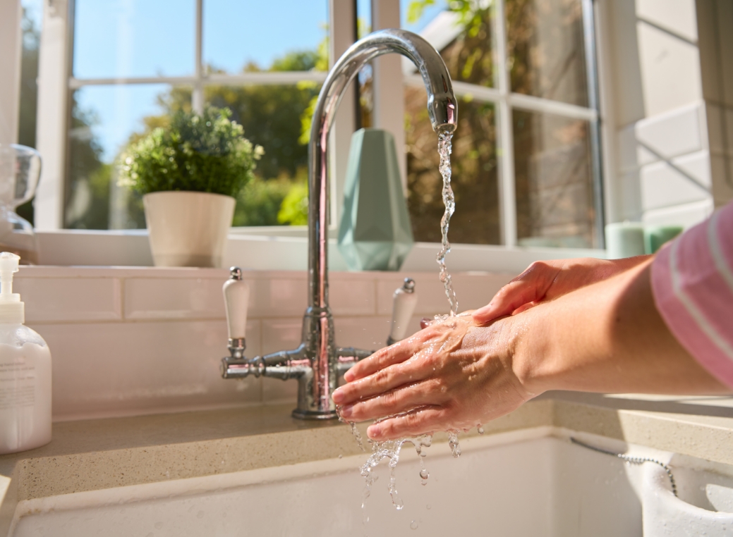 A pair of hands is shown close-up as water splashes from a faucet onto them. The image features a kitchen-style sink and faucet, with water coming from the faucet. The hands are placed under the water, and soap can be found on them as well as a pump on the left side of the sink. In the background of image, there is a large window and a potted plant that sits in front of it.