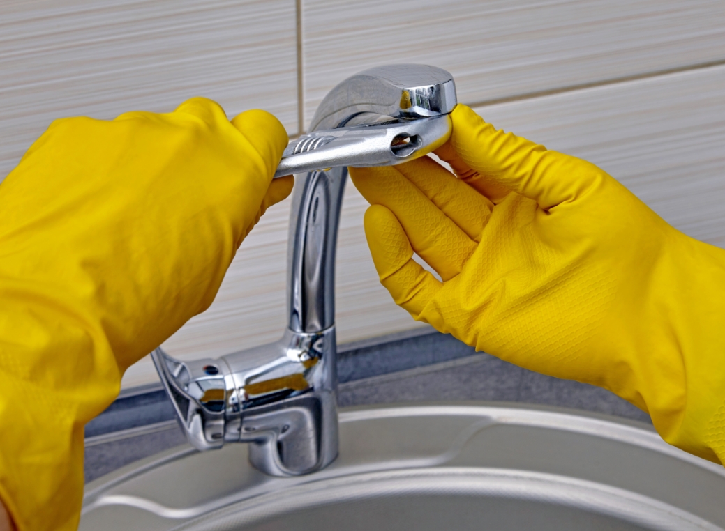A close-up, horizontal photograph shows a person’s hands wearing bright yellow rubber gloves while repairing a chrome kitchen faucet. The person is using a silver adjustable wrench in their left hand to tighten or loosen the base of the faucet, while their right hand holds the faucet steady. The faucet is mounted on a stainless steel double sink. The background features a neutral-toned, horizontally tiled wall, and the scene is brightly lit with a clean, practical focus on the task at hand.