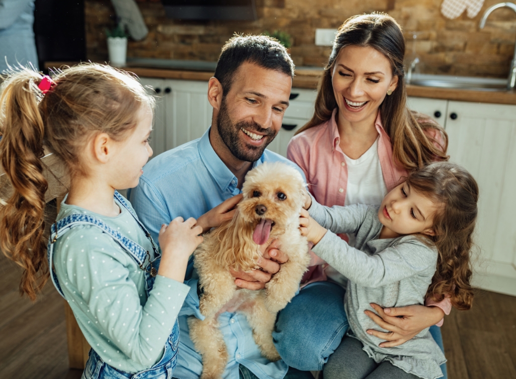 A family of four is gathered closely together in a bright kitchen, smiling and interacting with a small fluffy dog. In the center, a man wearing a light blue shirt holds the dog gently in his arms while smiling down at it. The dog, light brown in color, has its tongue slightly out and looks relaxed. To his left, a young girl with her hair tied back in a ponytail reaches out to pet the dog, focusing her attention on it. On the right side, another young girl leans in, also touching the dog with curiosity. Behind them, a woman in a pink shirt sits close, smiling warmly as she watches the children interact with the pet. The background shows a cozy kitchen with wooden cabinets, a tiled backsplash, and soft natural lighting that highlights the family’s expressions and the warm, homey atmosphere.